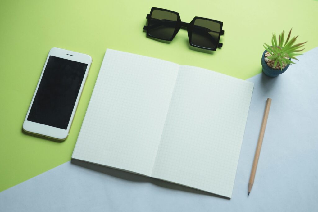 Flat lay of modern office essentials including smartphone, notepad, sunglasses, and plant.